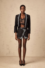 A woman wearing a black tweed jacket and matching skirt set with crystal embellishments The outfit features a tailored jacket paired with a short skirt both made from textured tweed fabric with sparkling crystal accents
