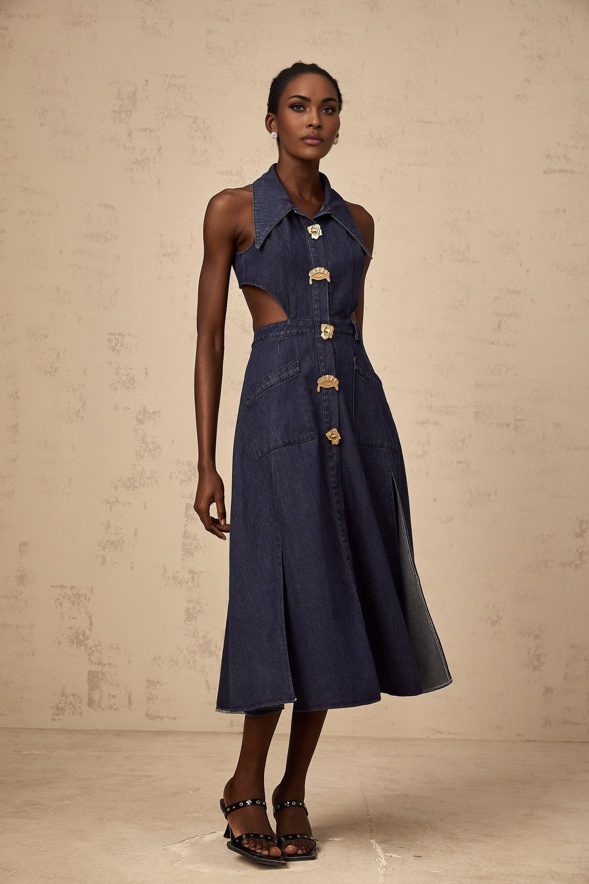 A woman wearing a dark blue denim midi dress with cutout details standing against a plain background