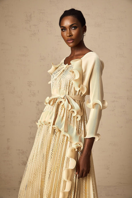 A woman wearing a neutralcolored midi dress with ruffled details and subtle gold accents standing in a relaxed pose