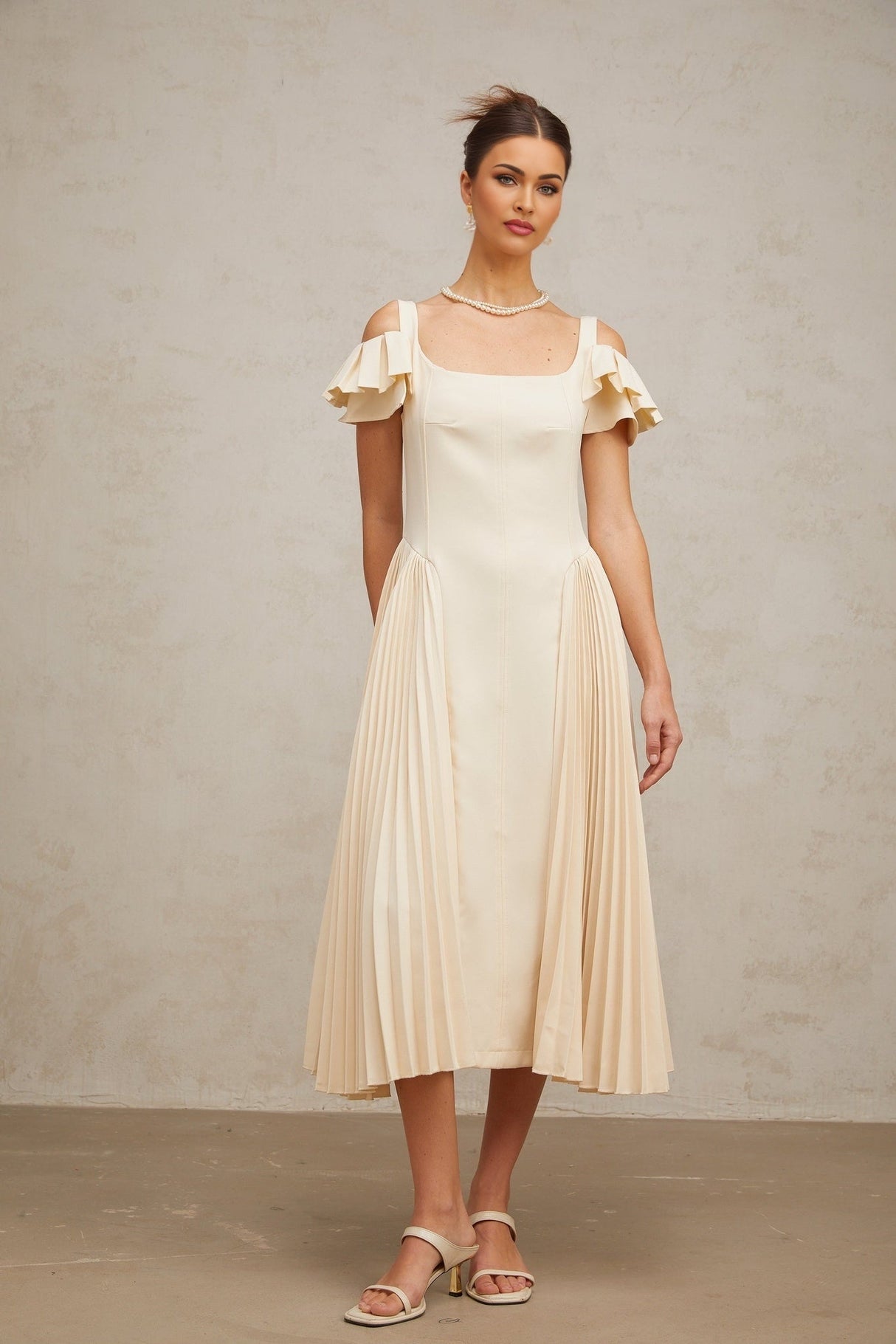 A woman wearing a neutralcolored pleated midi dress with a flared skirt and sleeveless design standing in a relaxed pose