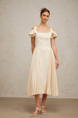 A woman wearing a neutralcolored pleated midi dress with a flared skirt and sleeveless design standing in a relaxed pose