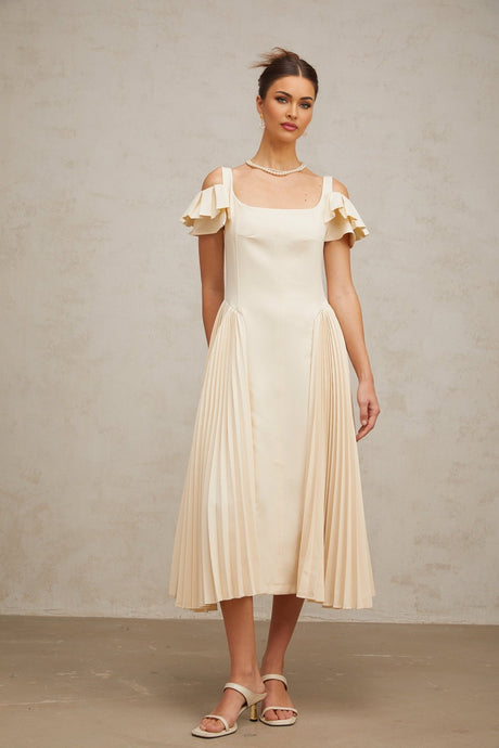 A woman wearing a neutralcolored pleated midi dress with a flared skirt and sleeveless design standing in a relaxed pose