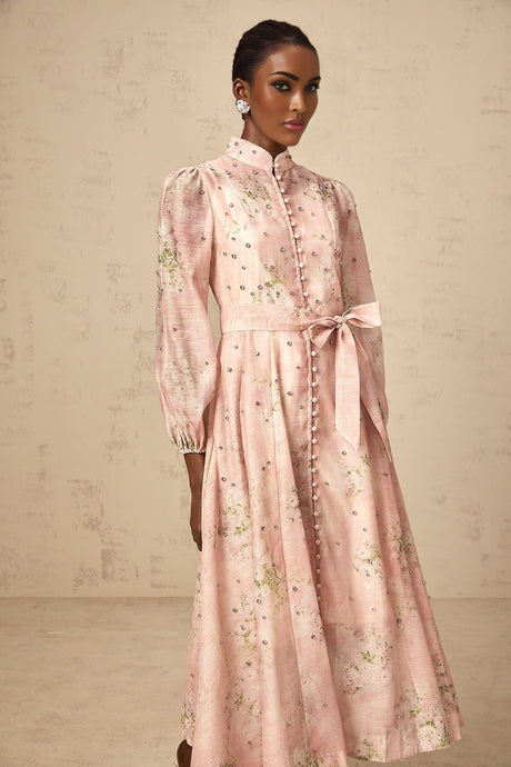A woman wearing a pink midi dress with floral print and embellishments standing against a plain background
