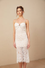 A woman wearing a white lace midi dress with a crystal embellished bustier top and delicate straps standing against a plain background