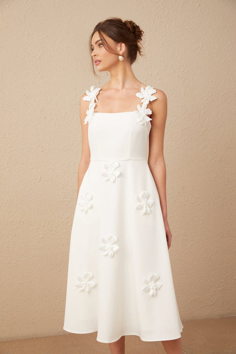 A woman wearing a white midi dress with a square neckline adorned with faux flower details standing against a plain background