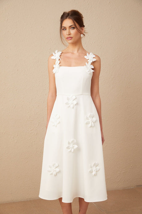 A woman wearing a white midi dress with a square neckline adorned with faux flower details standing in a neutral background