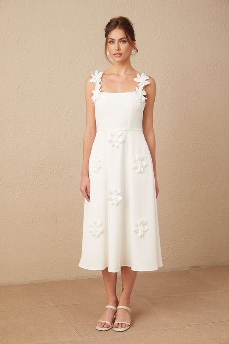 A woman wearing a white midi dress with a square neckline and faux flower embellishments standing against a plain background