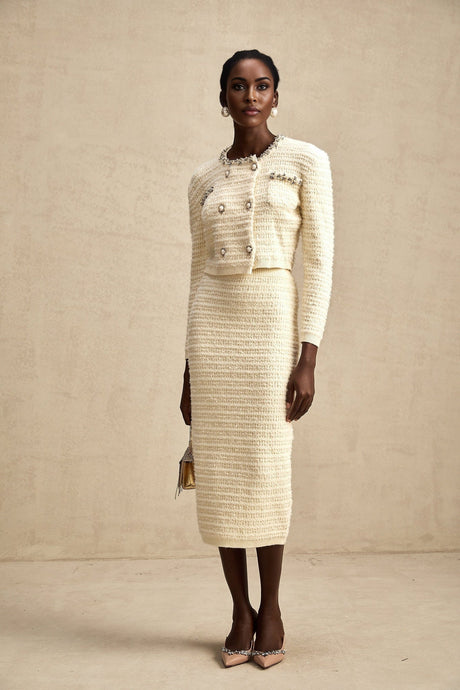 A young woman wearing a matching knit cardigan and skirt set with embellishments The outfit is light colored and features intricate detailing on both pieces