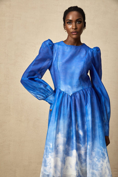 Woman wearing a blue gradient pleated midi dress with a flowing silhouette standing against a plain background