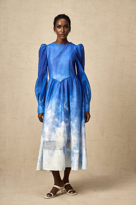 Woman wearing a blue gradient pleated midi dress with a flowing skirt and fitted bodice standing against a plain background