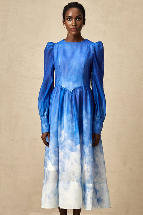 Woman wearing a blue gradient pleated midi dress with a flowing skirt and fitted bodice standing against a plain background