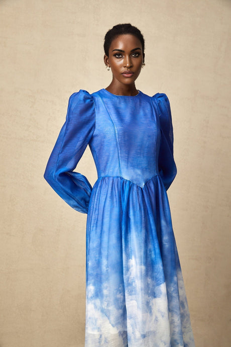 Woman wearing a blue gradient pleated midi dress with a flowy skirt and fitted bodice standing against a plain background