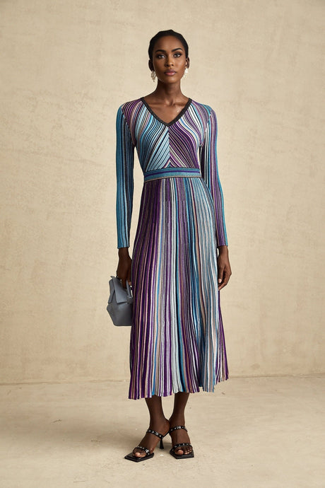 Woman wearing a blue metallic pleated midi dress with long sleeves and a cinched waist standing against a plain background