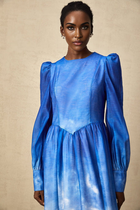 Woman wearing a blue midi dress with gradient color effect and pleated skirt standing against plain background