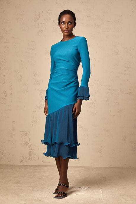 Woman wearing a blue midi dress with ruffled trim and panelled design standing in front of a plain background