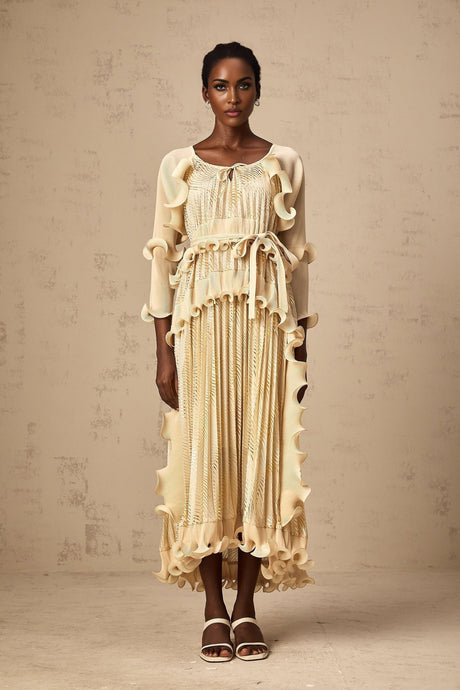Woman wearing a neutral toned midi dress with ruffled details and gold accents standing in a relaxed pose