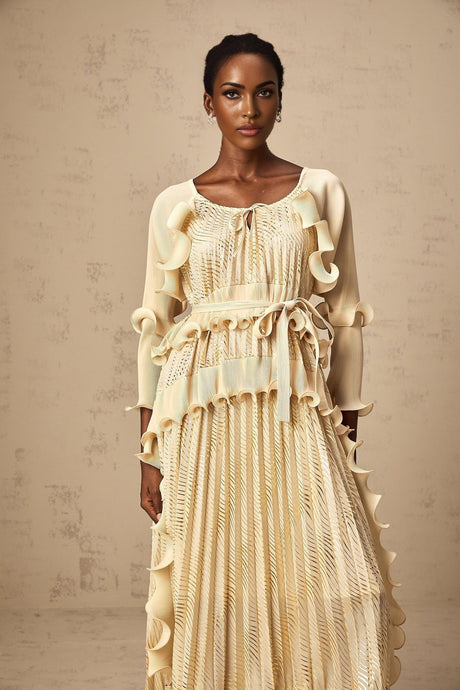 Woman wearing a neutralcolored midi dress with gilded accents and ruffled details standing in a relaxed pose