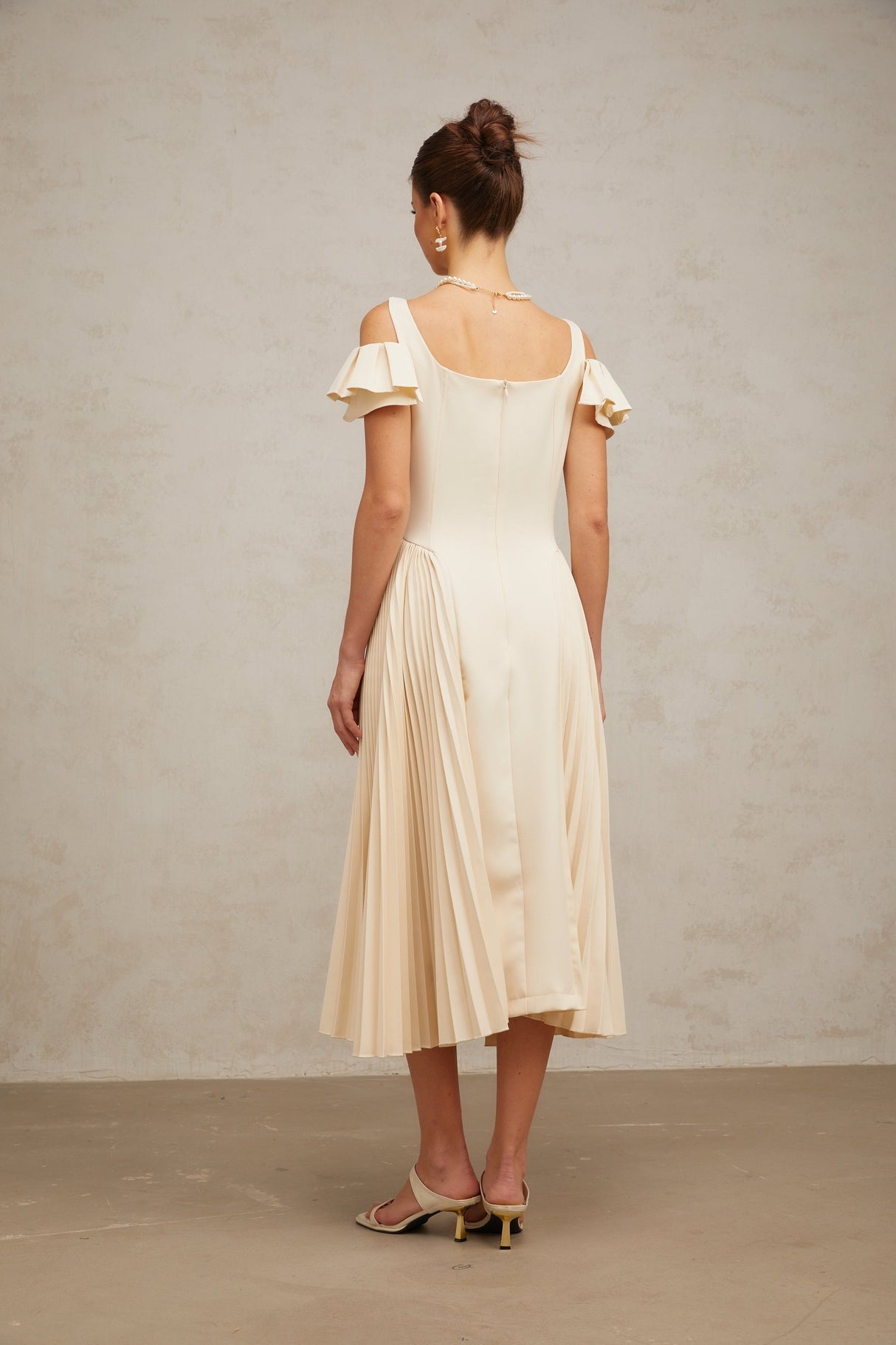 Woman wearing a neutralcolored pleated midi dress with a flared skirt and fitted bodice standing in a relaxed pose
