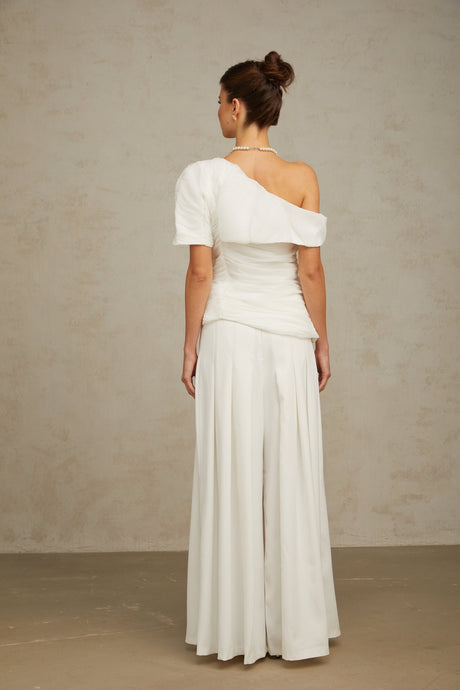 Woman wearing white matching set with creased top and trousers standing against plain background