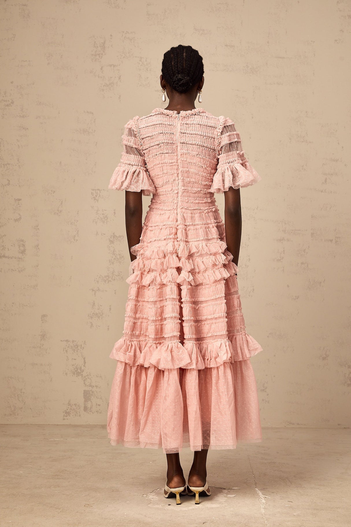 Young woman wearing a pink midi dress with dot details and ruffled tulle skirt standing in front of a plain background