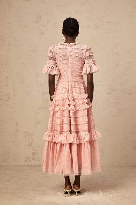 Young woman wearing a pink midi dress with dot details and ruffled tulle skirt standing in front of a plain background